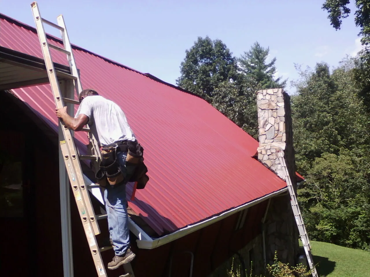Roofer on a ladder beside a red metal roof and stone chimney — Soffit & Fascia Repair by Roofing Brothers in Edgefield